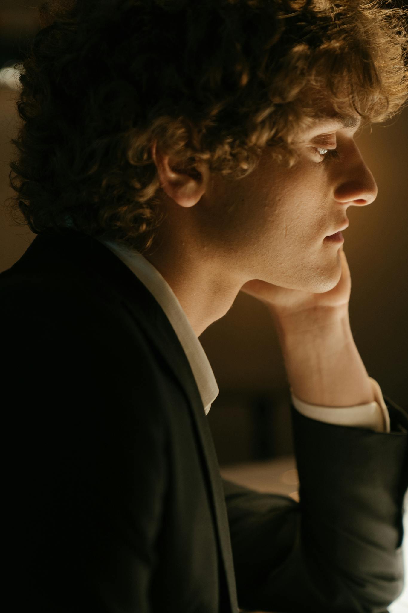 A thoughtful man with curly hair in a suit waits at a bar counter indoors.