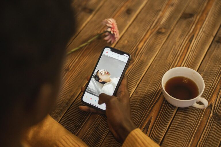 An individual using a dating app on a smartphone with a cup of tea nearby on a wooden table.